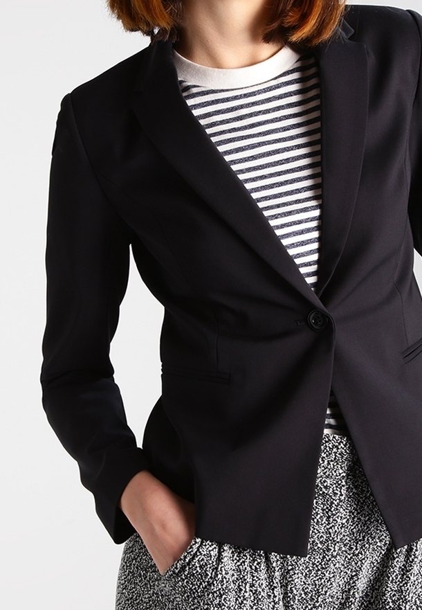 Frau in einem taillierten schwarzen Blazer über einem gestreiften Hemd, mit ihrer Hand in der Tasche einer gemusterten grau-weißen Hose.