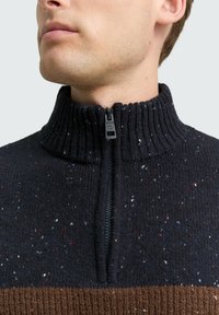 Nahaufnahme eines jungen Mannes, der einen schwarz gesprenkelten Strickpullover mit halbem Reißverschluss und hochgeschlossenem, geripptem Kragen trägt.