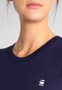 Lähikuva henkilöstä, joka pukeutunut tummansiniseen paitaan, jossa on pieni valkoinen "RAW"-logo rinnassa, näkyvissä osa kaulasta ja alahuulesta.