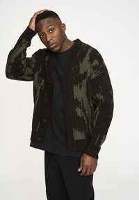 Dunkelgrüner und schwarzer gemusterter Cardigan mit Knopfleiste, gerippten Bündchen und strukturiertem Strick. Wird über einem schwarzen T-Shirt getragen.