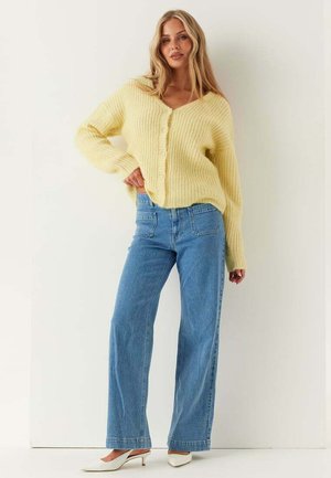 Kvinde stående iført en gul cardigan med knapper, blå jeans med høj talje og brede ben samt hvide spidse hæle mod en ensfarvet baggrund.