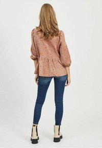 Femme aux longs cheveux portant un chemisier rose à fleurs, un jean skinny bleu et des bottines beiges, debout de dos sur un fond blanc.