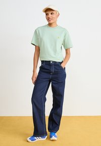 Ljust grön kortärmad t-shirt, mörkblå vida jeans i denim med vit söm, beige keps och blå träningsskor med vita ränder.