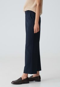 Pantalons taille haute bleu marine avec un design plissé, coupe ajustée, ourlets retroussés et une texture lisse, associés à des mocassins marron foncé.
