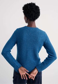 Rövid göndör hajú személy kék kötött pulóvert és sötét nadrágot visel, kezei összekulcsolva a háta mögött, világos háttér előtt áll.