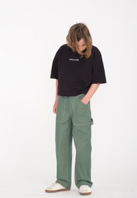 Czarna, oversize'owa koszulka z logiem "VOLCOM", zestawiona z zielonymi spodniami typu cargo o szerokich nogawkach, które mają liczne kieszenie oraz żółte przeszycia.