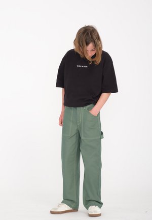 Czarna, oversize'owa koszulka z logiem "VOLCOM", zestawiona z zielonymi spodniami typu cargo o szerokich nogawkach, które mają liczne kieszenie oraz żółte przeszycia.