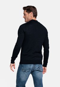 Donkerblauwe trui met lange mouwen, aansluitend ontwerp, gladde textuur en geribbelde manchetten, gedragen met lichtblauwe jeans. Zicht van achteren.