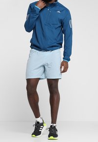 Chaqueta ligera azul con tejido texturizado, cierre frontal con cremallera y detalles reflectantes, combinada con pantalones cortos azul claro y zapatillas de correr negras con detalles neón.