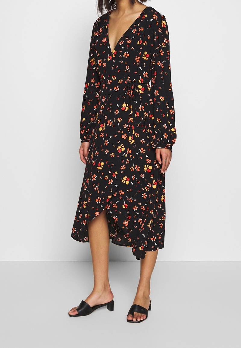 Femme portant une robe portefeuille noire à manches longues avec un imprimé floral rouge, orange et jaune, associée à des sandales noires à talons blocs et bout ouvert.