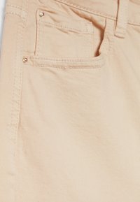 Primer plano de un bolsillo de unos vaqueros de mezclilla beige con costuras visibles y remaches de metal sobre la textura de la tela.