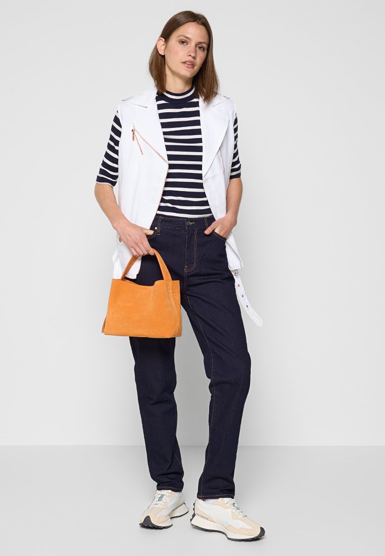 Canotta bianca con zip sopra un top a righe blu, jeans scuri e borsa arancione. Calzature sono sneakers di colore chiaro.