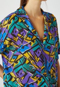 Camisa multicolorida con un audaz patrón abstracto en púrpura, azul, amarillo y negro, con cuello y mangas cortas.