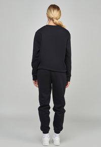 Mujer con cabello rubio en una coleta, vestida con sudadera negra y pantalones de chándal, y zapatillas blancas, de pie de espaldas sobre un fondo liso.