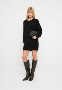 Zwarte gebreide sweaterjurk met kabelpatroon, lange mouwen en geribbelde randen. Gecombineerd met bruine leren laarzen tot aan de knie en een kleine zwarte tas.