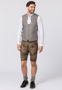 Graue, strukturierte Weste kombiniert mit braunen Lederhosen, die aufwendige Stickereien aufweisen, knielang sind und dunkle Schuhe mit grauen Socken tragen.
