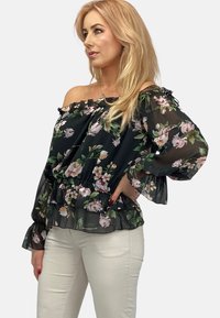 Czarna bluzka off-shoulder z kwiatowym wzorem, z falbaniastymi rękawami i dołem. Przezroczysty materiał z różowymi i zielonymi kwiatami. W zestawie z jasnymi spodniami.