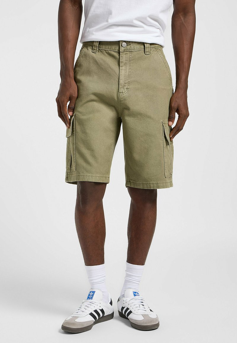 Shorts cargo vert olive avec deux poches latérales, fabriqués en tissu résistant. Dotés d'une fermeture à bouton et de passants de ceinture. Portés avec des chaussettes blanches et des baskets.