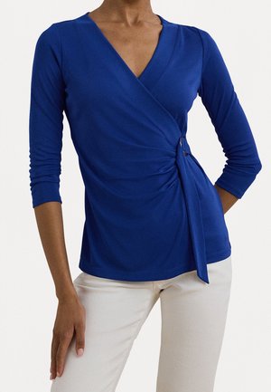 Femme portant un haut cache-cœur à manches longues bleu royal avec un nœud sur le côté et un pantalon blanc, visible du cou jusqu'aux hanches sur un fond blanc.