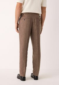 Pantalones tailored marrones con un acabado texturizado, que presentan bolsillos traseros con botones negros, corte recto y un ligero estrechamiento en los tobillos.