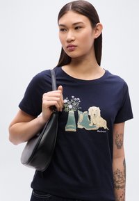 Μπλε βαμβακερό t-shirt με σχέδιο που απεικονίζει πράσινες μπότες, λουλούδια και ένα μπεζ σκύλο. Το μοντέλο κρατάει μια μαύρη τσάντα μέσης.