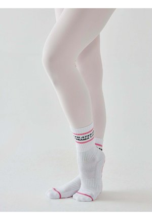 Nogi w białych rajstopach ze skrzyżowanymi stopami, ubrane w białe skarpetki z różowymi paskami i czarnym tekstem, stojące na jednolitym jasnym tle.