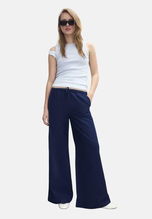 Kobieta ubrana w czarne szerokie spodnie, białą asymetryczną bezrękawnikową bluzkę, białe buty i czarne okulary przeciwsłoneczne, stojąca z jedną ręką w kieszeni.