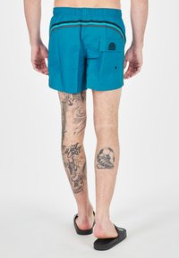 Costume da bagno turchese da uomo con vita elastica, strisce nere a contrasto e tasca posteriore. Indossato con sandali neri, con tatuaggi sulle gambe.