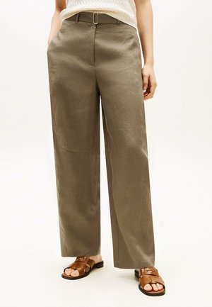 Person trägt braune, hoch taillierte, weit geschnittene Hose mit Gürtel und braunen Sandalen mit offenen Zehen und steht mit entspannten Händen an den Seiten.