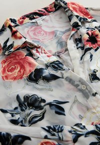 Blouse transparente avec motifs floraux en velours brodés en fils rouge, noir et or sur tissu blanc, avec col et fermeture par boutons.
