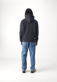Person with long dark hair wearing a black hoodie, blue jeans, and sneakers, standing facing away against a plain white background.
