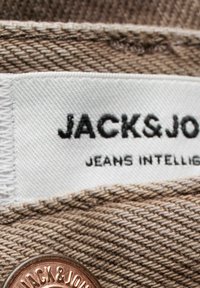Gros plan sur un tissu en jean beige avec une étiquette blanche Jack & Jones et un bouton couleur cuivre avec le nom de la marque en relief.