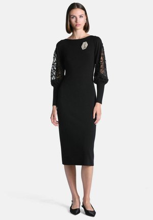 Femme portant une robe midi noire ajustée avec des manches bouffantes en dentelle, une broche géométrique sur la poitrine et des talons noirs à bout pointu.