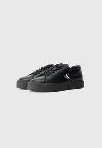 Adidași negri din piele, cu o textură netedă, talpă din cauciuc negru plat și logo brodat alb pe lateral. Design cu șireturi și vârf jos.