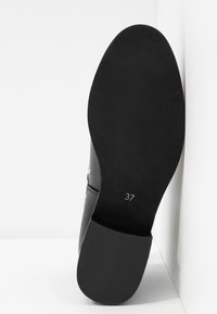 Suela de zapato negra mirando hacia adelante con el número de talla 37 impreso cerca del talón, posicionada de pie contra un fondo blanco en la esquina.