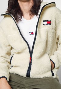 Chaqueta de forro polar color crema con ribete azul marino, cierre completo y tirador rojo. Cuenta con dos parches de marca en el pecho y una textura suave.