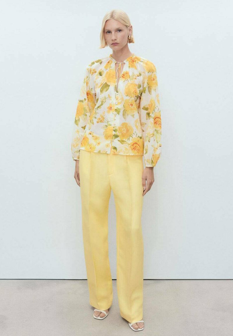 Blumenbluse mit gelben und orangen Rosen, Knopfverschluss vorne, geraffter Ausschnitt. Kombiniert mit hoch taillierten gelben Hosen aus einem glatten Stoff.