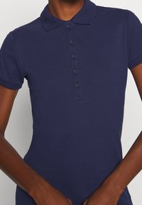 Polo de manga corta azul oscuro con una tapeta de cinco botones, llevado por una persona de piel oscura, con los brazos relajados a los lados.