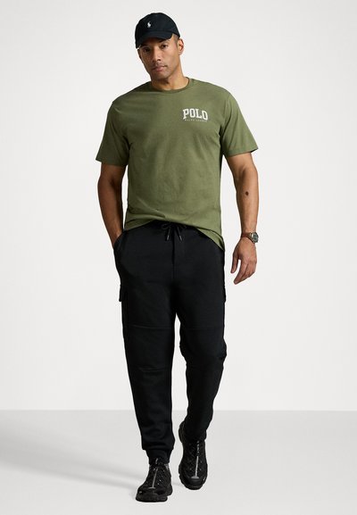 Olive spalvos medvilnės marškinėliai su "POLD" logotipu; juodos krovinių kelnės su elastingu juosmeniu; juodi kalnų batai; klasikinė juoda kepurė.