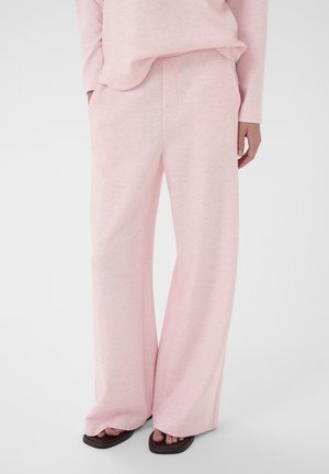 Osoba v nosení voľných svetloružových nohavíc a zodpovedajúceho topu, stojaca s jednou rukou v vrecku, obutá v tmavohnedých sandáloch s otvorenou špičkou.