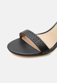 Ανοιχτό μαύρο πέδιλο με δερμάτινο λουράκι με τρύπες μπροστά και λεία σόλα, εμφανιζόμενο σε απλό λευκό φόντο.