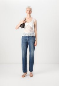 Hvit blondebluse med korte ermer, kombinert med lyseblå rette jeans. Modellen holder en liten mørk håndveske og har på seg brune sandaler med stropper.