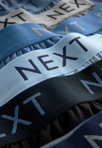 Textilné pásy v odtieňoch modrej, šedej a čiernej, s nápisom "NEXT" v kontrastných vyšívaných písmenách a volánovou textúrou.