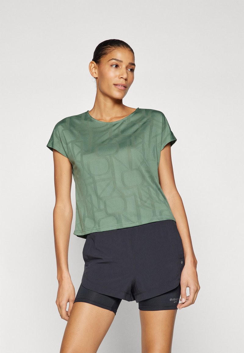 Vihreä lyhythihainen crop-top, jossa on geometrinen kuvio, yhdistetty mustiin urheilushortseihin. Kangas näyttää sileältä ja kevyeltä.
