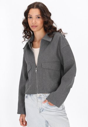 Giacca grigia cropped in morbido tessuto con colletto, zip frontale e due tasche sul petto, abbinata a jeans azzurri chiari.