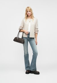 Kashmir kardiganis vaale bež boje, bijela majica, svijetloplave uske traperice, i smeđe platforme; drži tamnosivu torbicu.