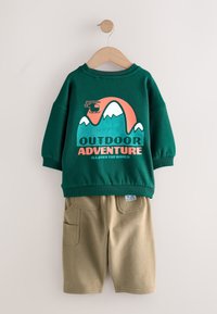 Chemise verte à manches longues avec illustration de montagne et d’hélicoptère et texte « Outdoor Adventure », accompagnée d’un short cargo beige sur un cintre.