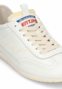 Baskets en cuir blanc avec des lacets beiges, une couture à bord festonné, et une étiquette en tissu bleu et rouge sur la languette portant l'inscription "ENTJPN".