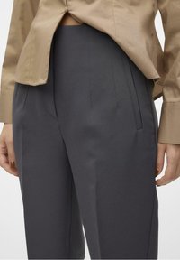 Dopasowane ciemnoszare spodnie o gładkiej fakturze, z przodu z zakładkami i bocznymi kieszeniami, w połączeniu z jasnobeżową bluzką.