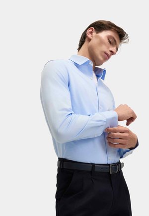 Camicia azzurra chiara con maniche lunghe, colletto a bottoni e texture leggera, indossata con pantaloni neri e una cintura nera.
